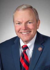 Photo of Ohio State Senator Bill Reineke wearing a navy blue jacket with a red tie smiling in front of a gray background